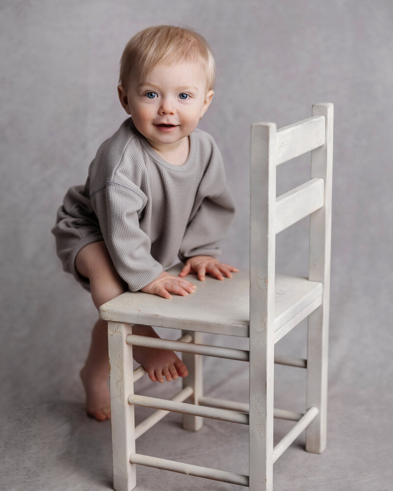 Baby boy climbs white toddler chair in Murrysville portrait studio.