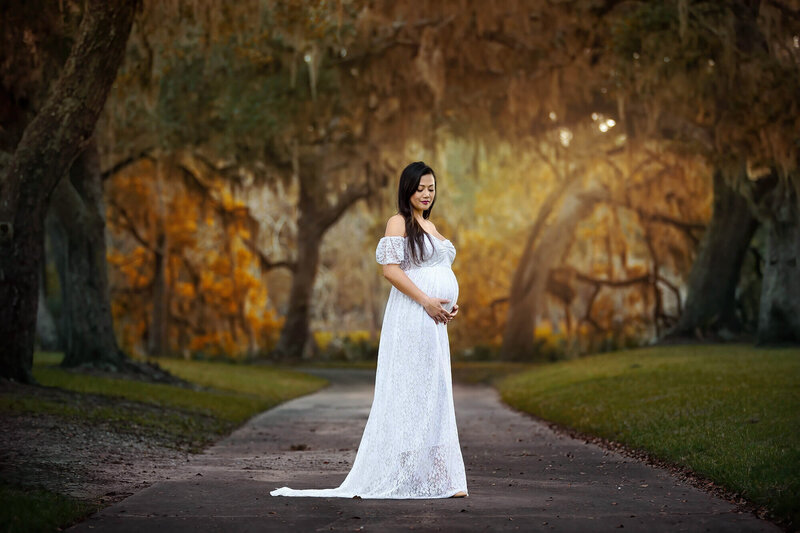 Beautiful expecting mama wearing a white dress holding her belly in front of beautiful trees  in Houston, Texas.