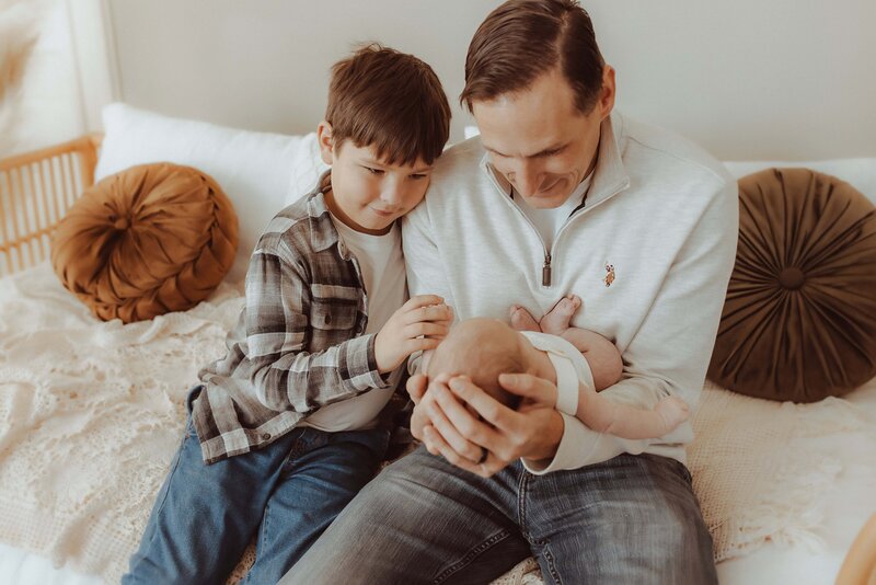 A man holds a baby while a young boy beside him looks on, both sitting on a bed with decorative pillows.