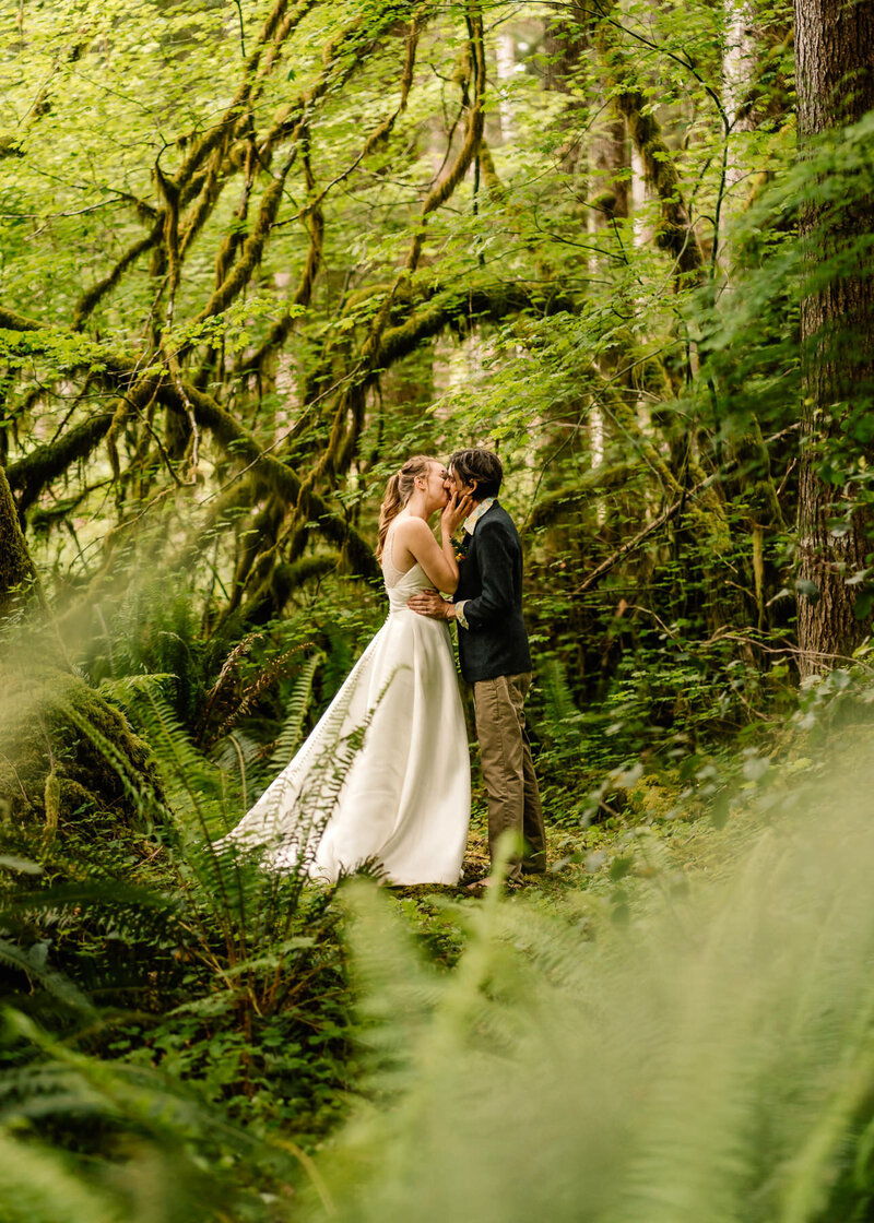after learning how to elope in Washington State, a couple in wedding attire kisses in the rain forest. They are surrounded by lush foliage
