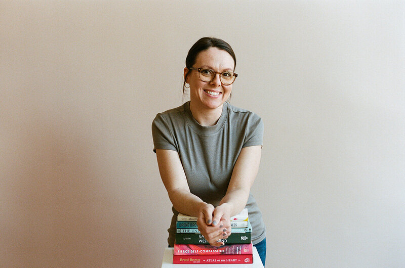 A woman with glasses, Kristen Simons, sits beside a tall stack of books, smiling and engaged in reading.