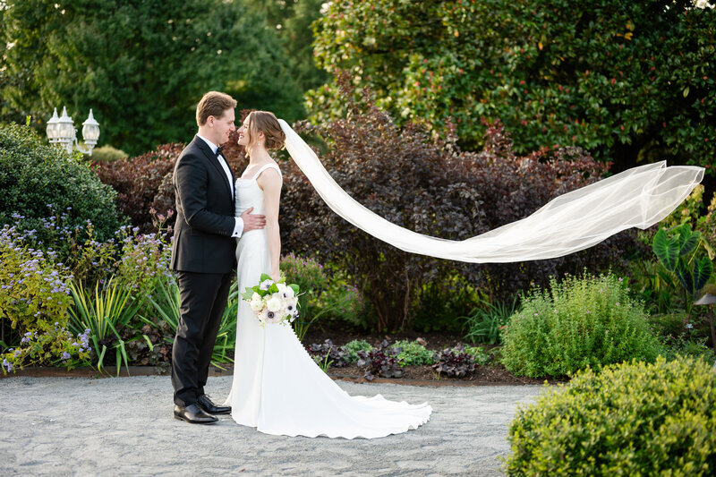 A bride and groom look at one another while the bride's veil floats behind her outdoors