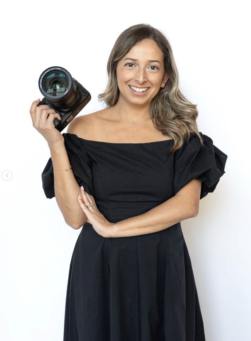 Portrait of Andrea Silva, luxury wedding photographer, wearing an off-shoulder black dress and holding camera