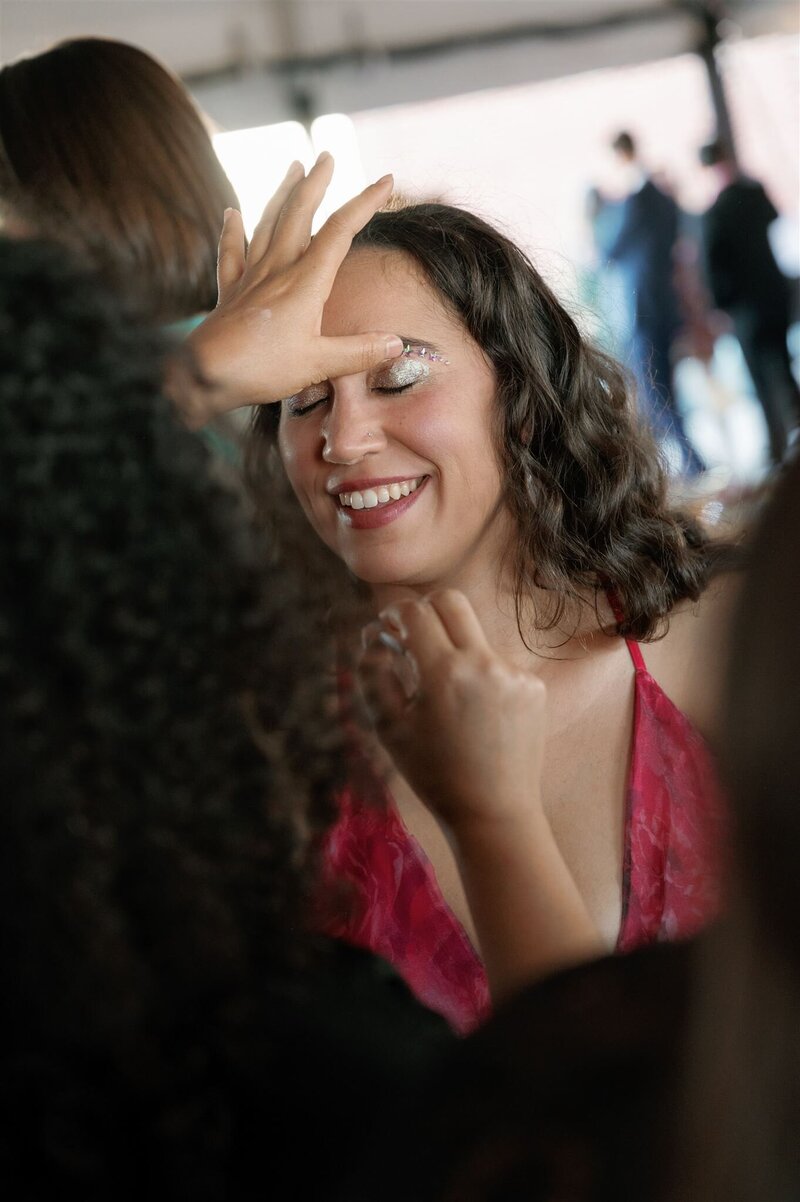 Wedding guest sitting with eyes closed in a hot pink halter dress receiving glitter eye shadow from a rave makeup station at a wedding on the terrace at the Line Hotel DC