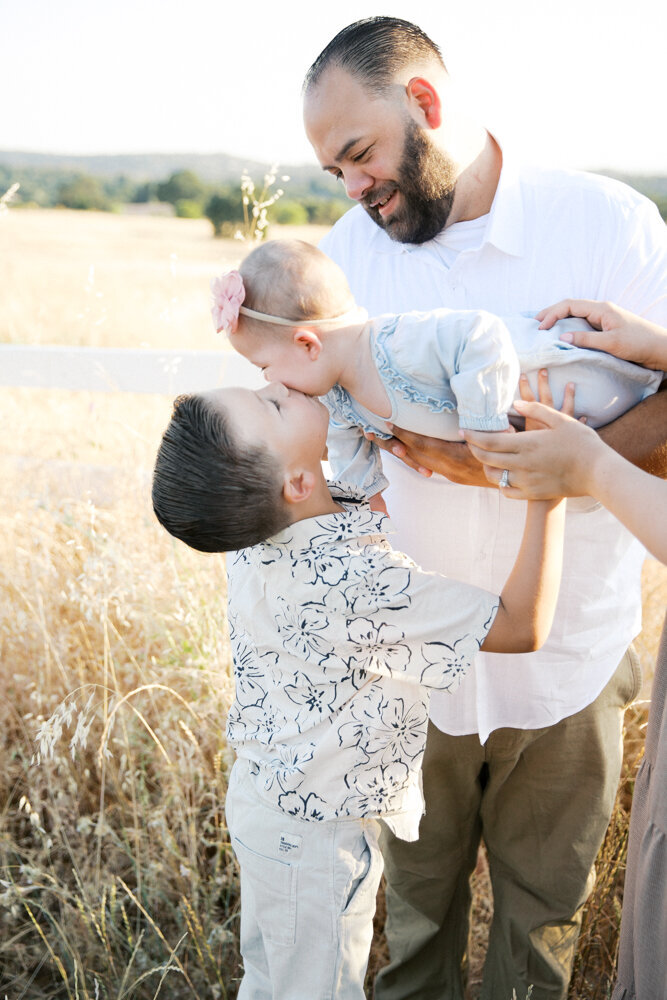 Image of big brother reaching up to kiss his baby sister in dad's arms,  taken by Sacramento Newborn Photographer Kelsey Krall