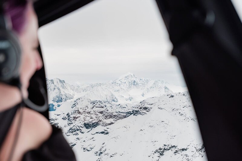 A bride in formalwear looks out of the helicopter window during their elopement wedding on an Alaska Glacier.  the mountains are crisp in focus with bright white snow covering the craggy peeks.    Her headset to be able to hear the pilot is visible at the edge of the frame, and you can see she's smiling. 