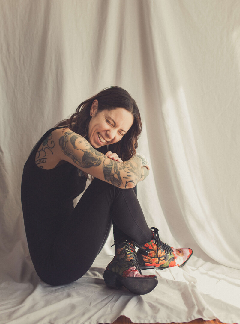 tattooed girl wearing black & bright embroidered boots is laughing sitting on simple white backdrop