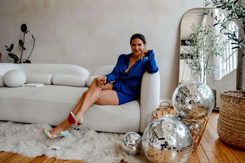 Emily Michele sitting gracefully on a cream sofa in a royal blue outfit beside disco balls, embodying power, confidence, and celebration.