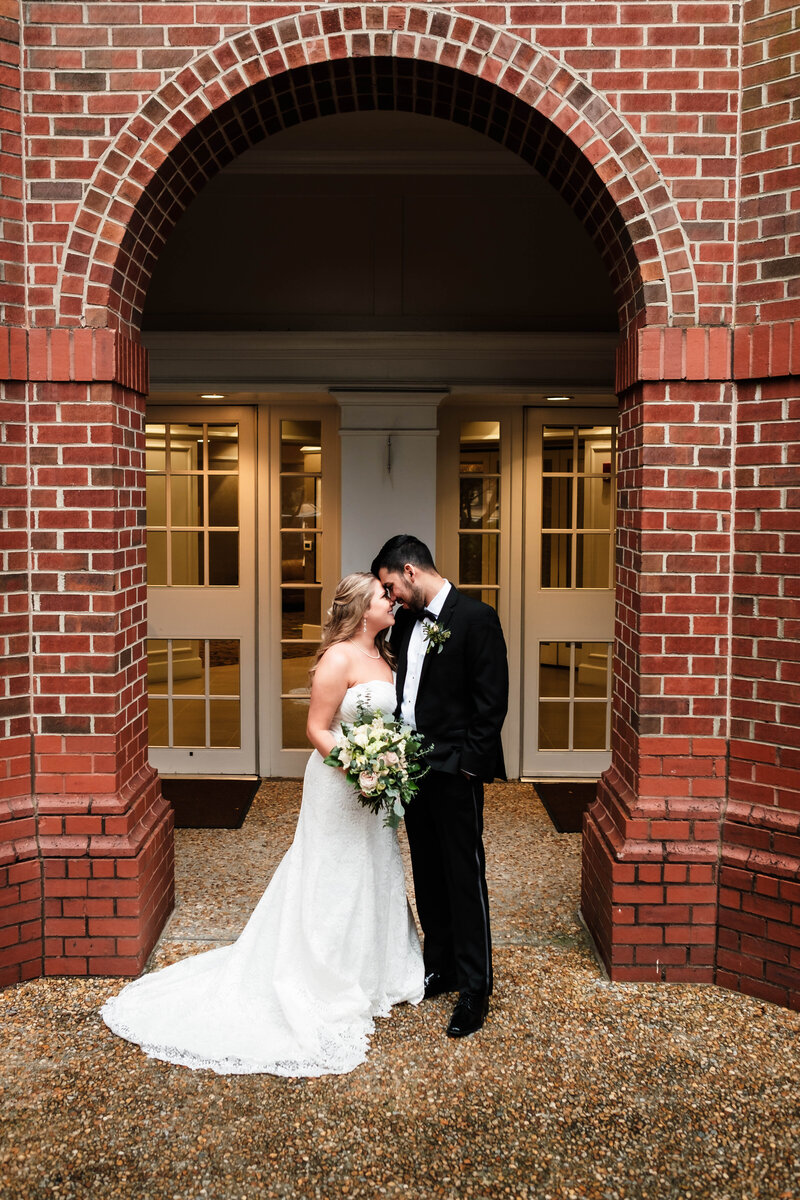 A bride and groom share an intimate moment under the elegant brick archway of the Auburn University Hotel. The bride holds a lush bouquet as they lean in close, capturing the romance and sophistication of their wedding day. This timeless image showcases the artistry of Alabama wedding photography in a classic Auburn setting.