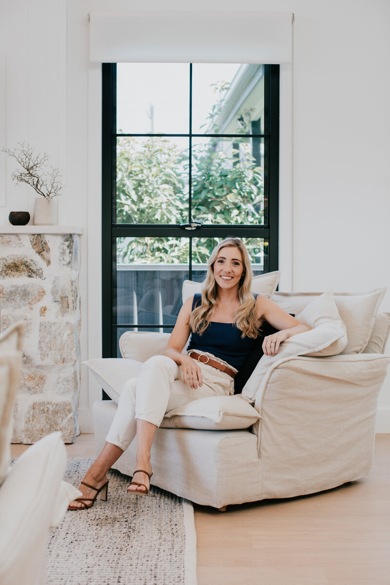 A woman relaxes in a cozy armchair by a large window, enjoying the bright and airy room with neutral decor, crafted through full-service interior design in Newcastle.