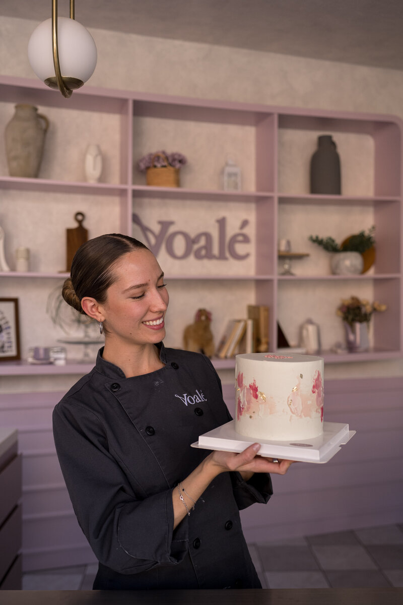 Voalé Bakery brand identity showcased in lifestyle photo of pastry chef holding floral cake inside branded pastel interior.