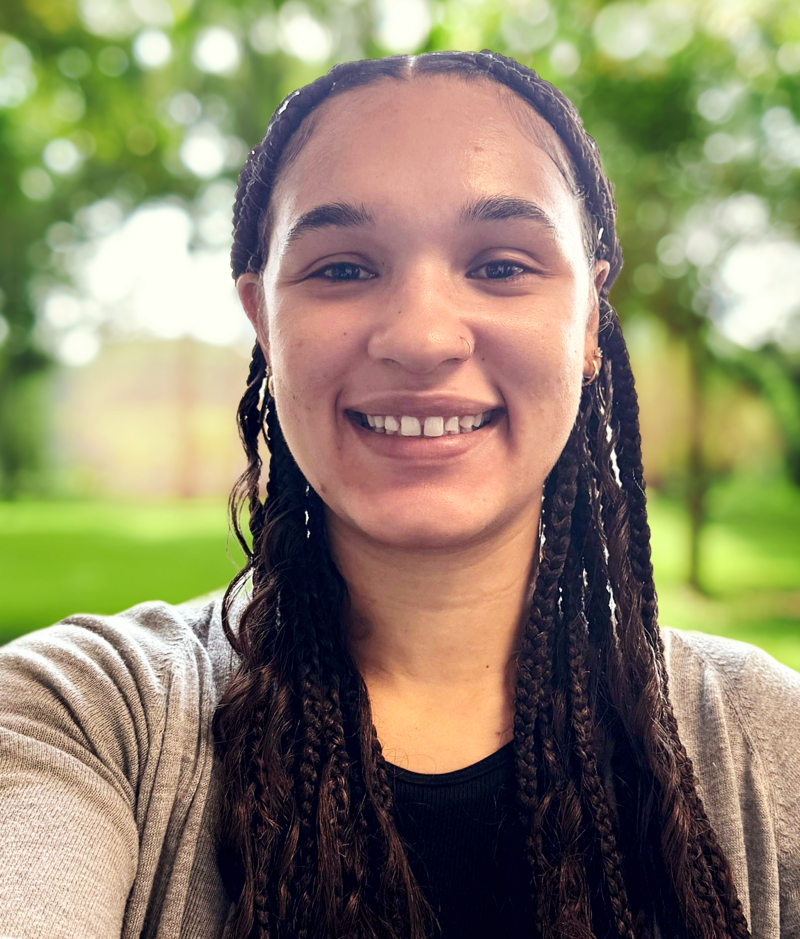 Haili is a smiling young woman with long dark braided curls, dark brown eyes, wearing a black shirt with a cream colored cardigan. 