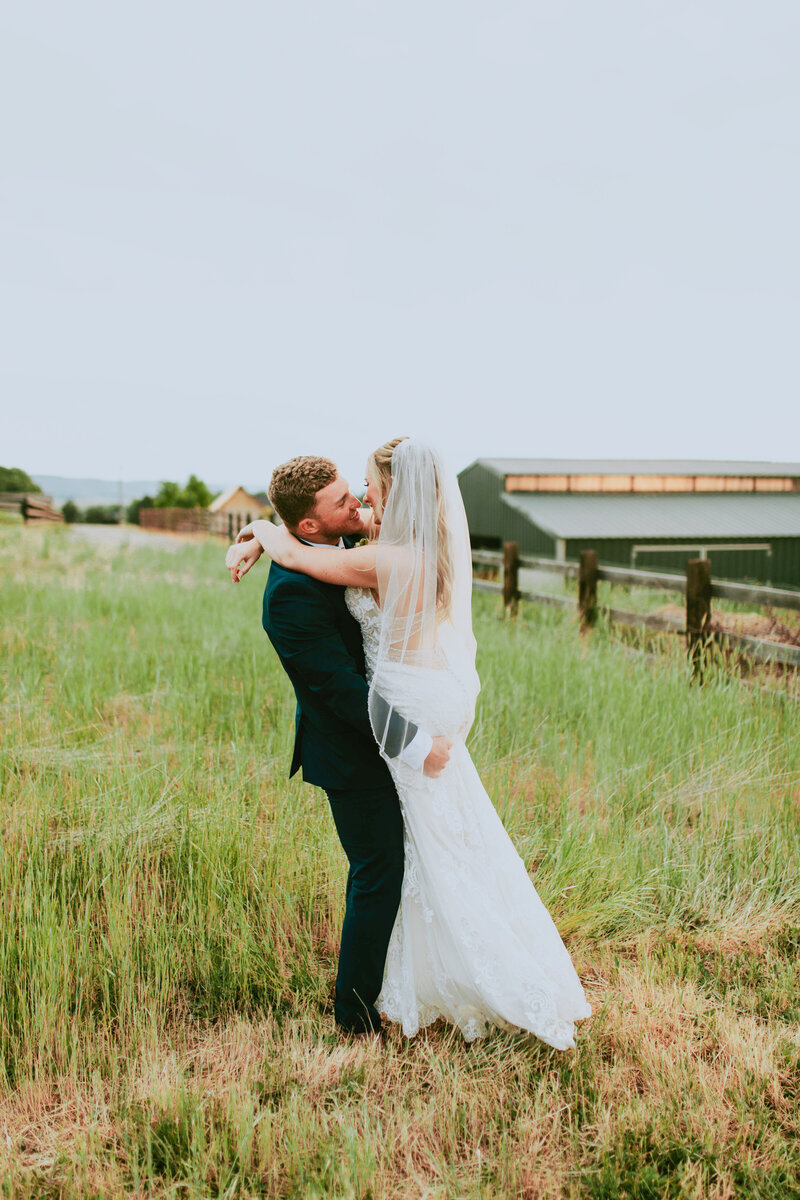 Groom lifting his wife up for a kiss out in the pasture.