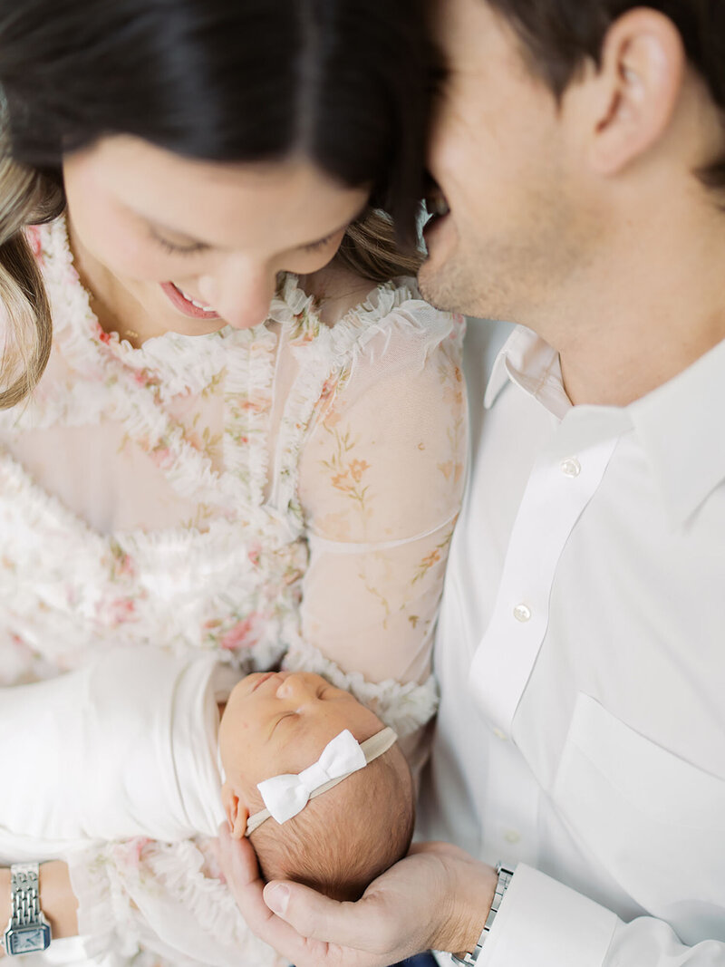 A mother and father are close together, holding their newborn baby. The father is whispering in his wife's ear while she smiles lovingly at their baby, capturing a tender and intimate family moment. Photo by a Austin Newborn Photographer
