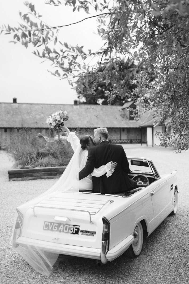 A couple, dressed in a wedding gown and suit, sit happily on the back of a vintage convertible at The Farmhouse at Redcoats. The bride holds up a bouquet as they pose in a gravel courtyard with rustic buildings in the background. The black-and-white image captures their timeless joy.