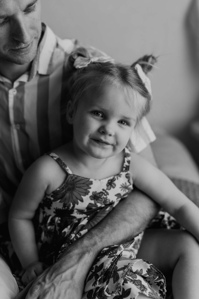 A toddler girl smiles while being held in her daddy's big arms. She is wearing a floral sleeveless dress with her hair in adorable little buns on top of her head.