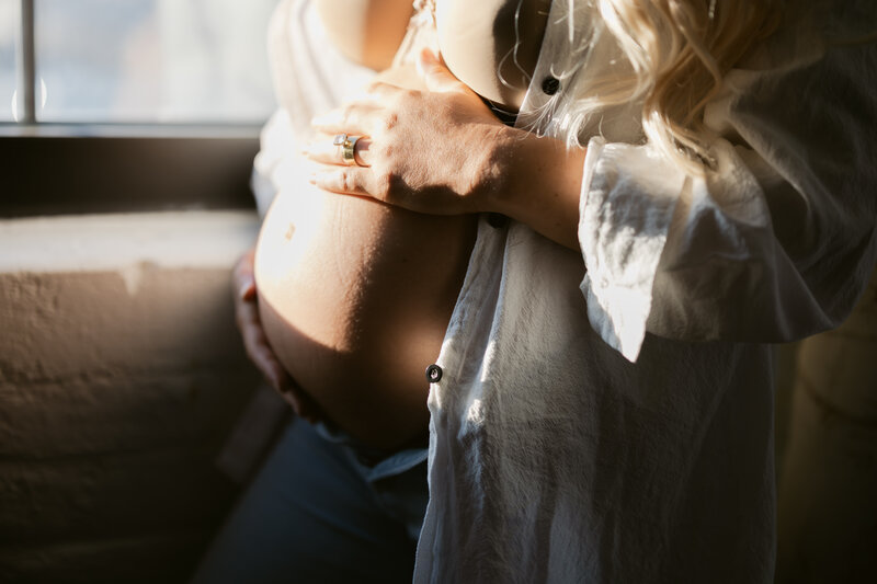 an expecting mother holds her belly as she stands in a window with golden sunlight hitting her belly. She wears an open white collared shirt and nude bra