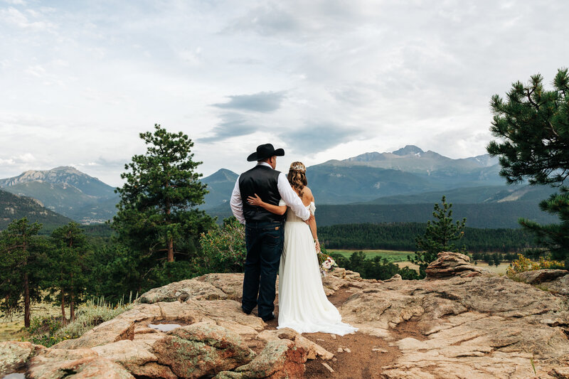 a couple is standing on rocks looking at a mountain view. the man is wearing a cowboy hat