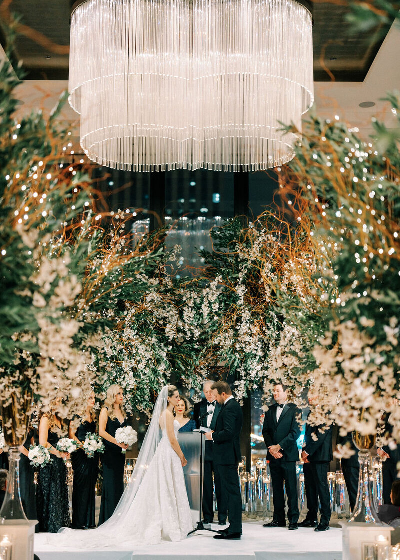 A lively wedding ceremony at The Langham Chicago with guests seated around a beautifully decorated aisle with dense floral arches and a chandelier made of greenery and white blooms.