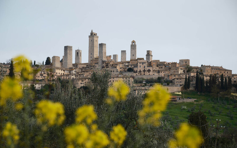A scenic view of San Gimignano, an Italian village with medieval stone towers and historic buildings, set against a cloudy sky. Yellow flowers and greenery add vibrancy to the scene, reminiscent of Caribbean getaways with their lush landscapes and unique charm.