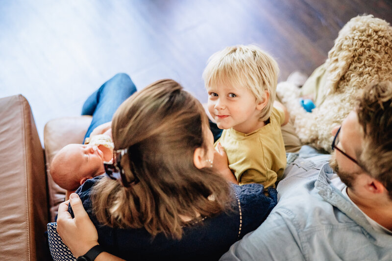 A toddler sitting with their parents and newborn sibling, smiling at the camera while the baby sleeps peacefully.