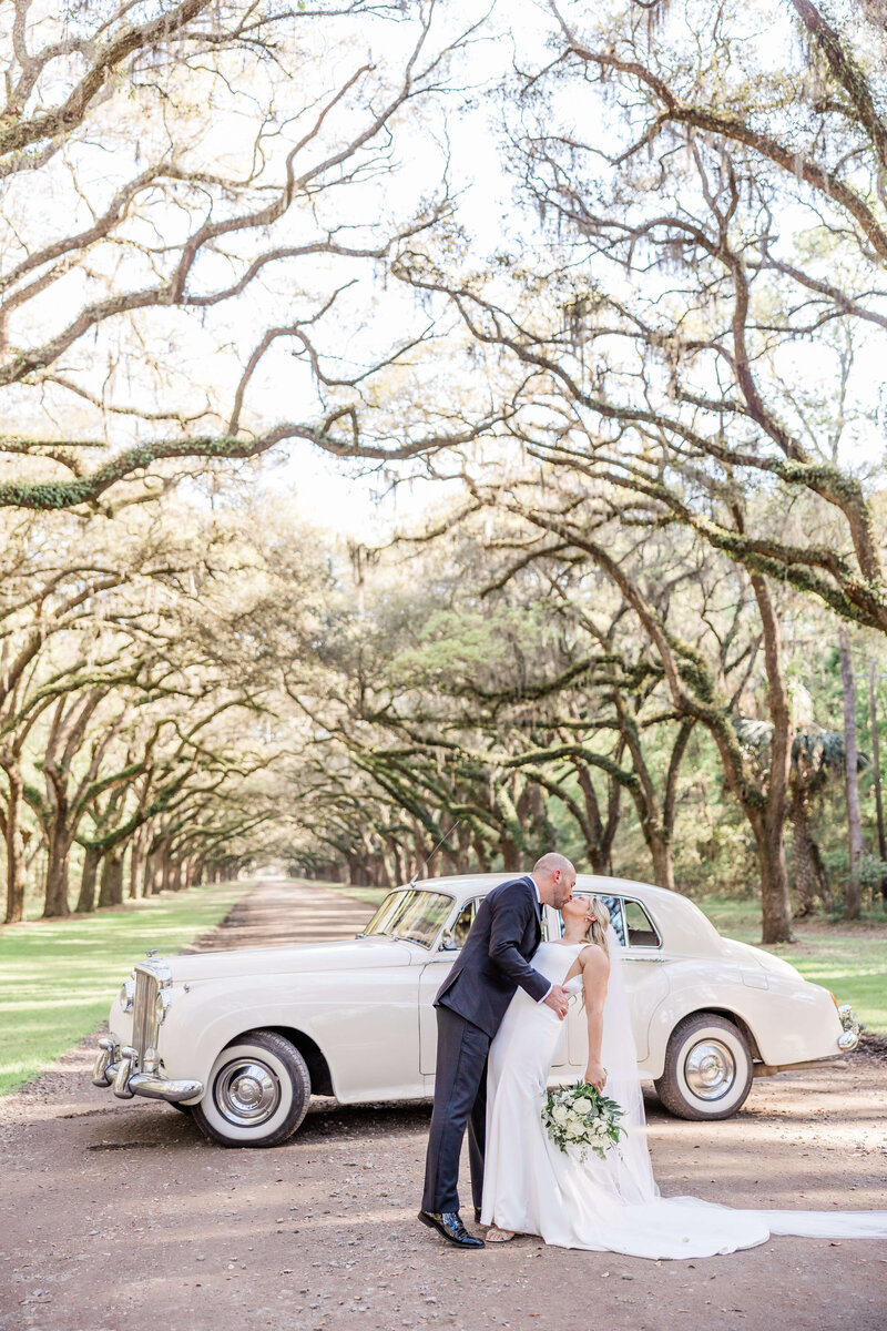 Bride and groom sharing a kiss beside a vintage white car under the moss-draped oak avenue at Wormsloe Historic Site in Savannah, GA