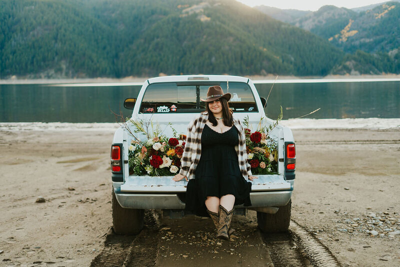 Grand Tetons senior session. A girl is sitting on the bed of a truck that has florals filling the back. She's wearing cowboy boots and a cowboy hat and softly smiling at the camera.