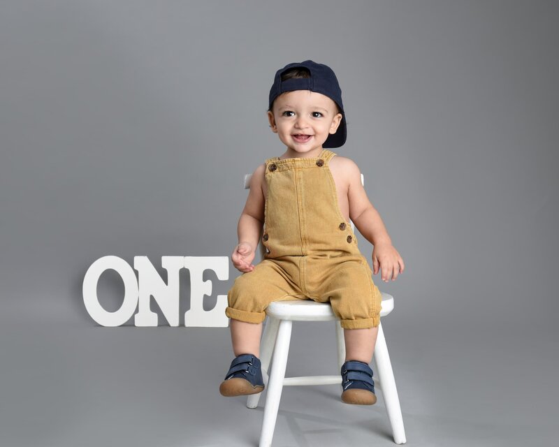 A baby boy wearing yellow overalls and a backward cap, sitting on a white chair with a cheerful expression, next to a "ONE" sign in a minimalist gray background setup.