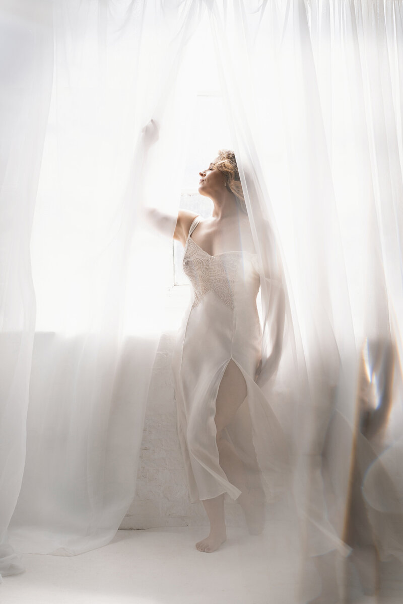 Light and airy boudoir photograph of a woman standing in between curtains, shot by Carrie Roseman, Connecticut