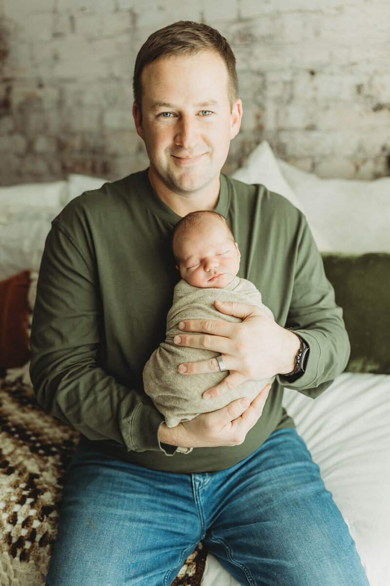 dad with blue eyes in an olive green henley holding his swaddled baby boy in harrisburg newborn photos