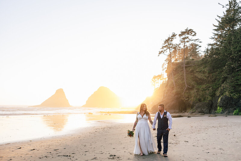 A bride and groom walk hand in hand on the beach as the sun sets behind them in Oregon.