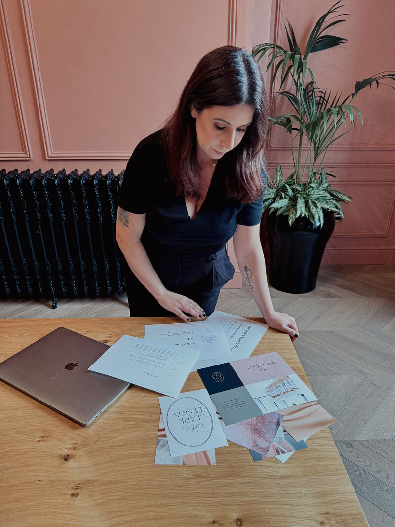 Roberta Rechena from By Roberta Design lays out design sheets on a table in front of her as she examines them. She wears a short sleeved black top and trousers