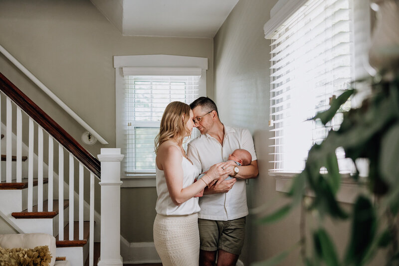 A mother and father holding infant in their home with stairs in the background and plants in the foreground