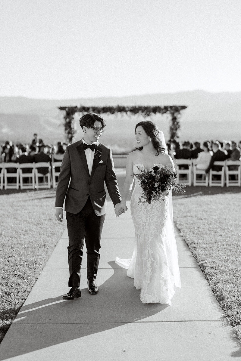 Captured in stunning black and white, this outdoor California wedding showcases the couple’s joyful walk down the aisle, framed by the distant mountains and a beautifully adorned floral arch. The bride’s lace gown and the groom’s tailored suit perfectly complement the elegant, natural setting.