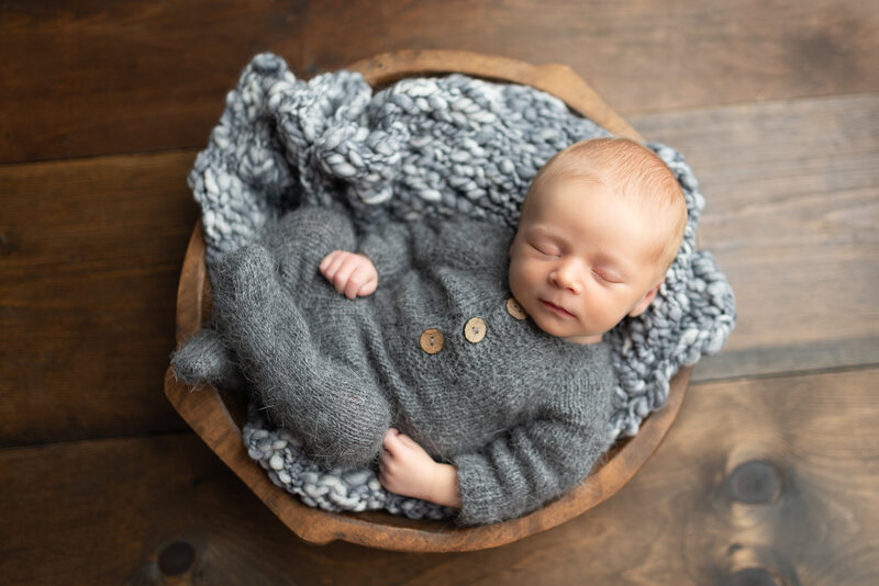 Newborn boy in grey romper with wooden buttons, curled in a rustic wooden bowl with textured grey knits – captured from overhead.
