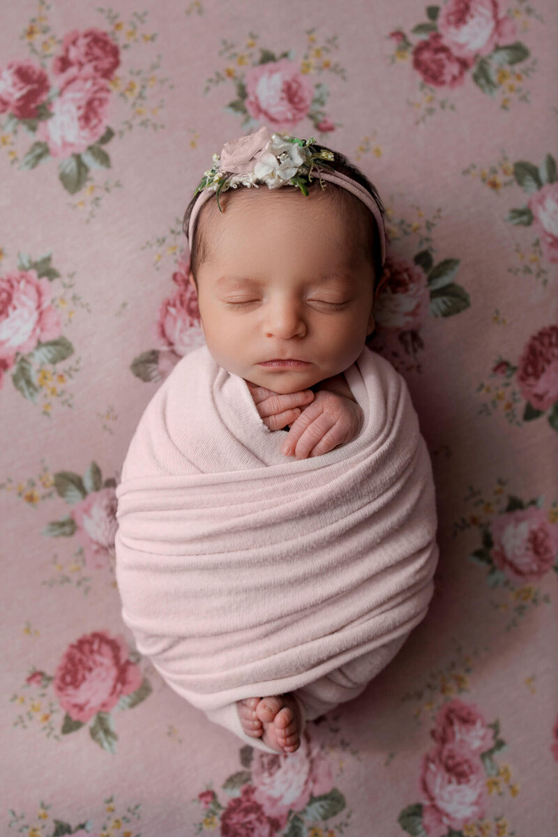 A newborn baby girl peacefully sleeps swaddled in a light pink wrap, adorned with a floral headband, lying on a floral-patterned background.