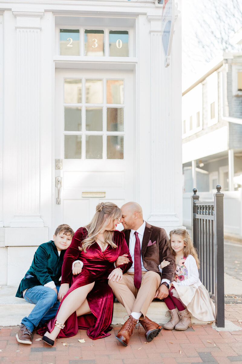 A mom and dad sit on some city steps with their young son and daughter hugging their arms in Christmas colors