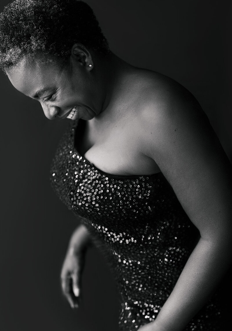 Black & white image of a lady in a sequin dress looking at the floor and smiling. Taken during a 40 over 40 photoshoot by Norwich Portrait Photographer Claire Howes, Norwich.