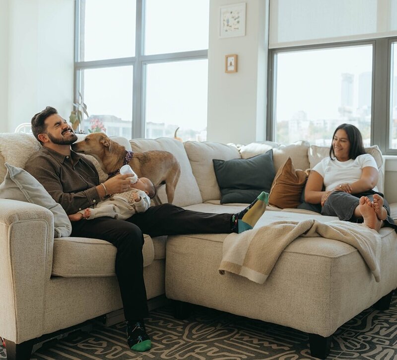 father feeding baby with dog and wife looking on during in home newborn photos captured by NYC newborn photographer Elsie Goodman 