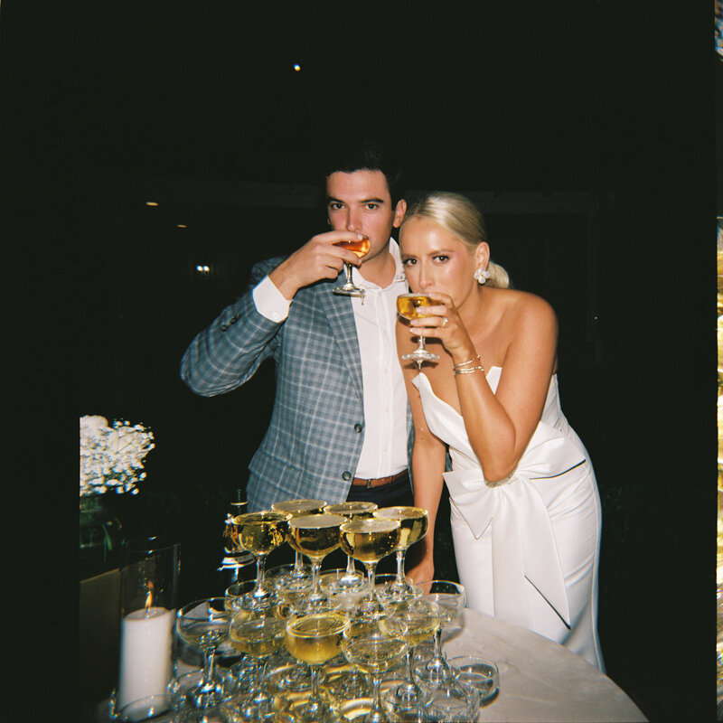 Flash film photo of bride and groom drinking glasses from their champagne tower