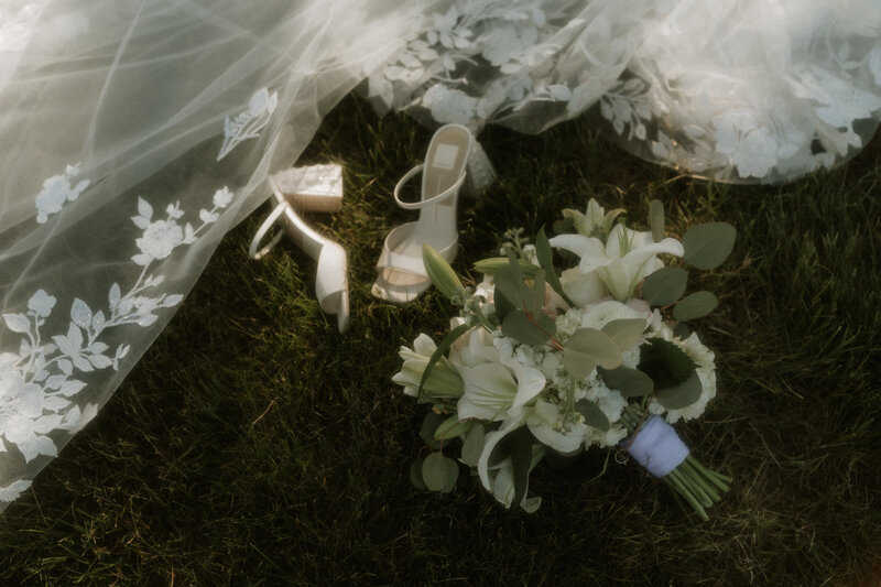 Bridal bouquet, heels, and lace veil resting in the grass, captured by Rainstorm Photo & Video in soft morning light with romantic, cinematic tones.