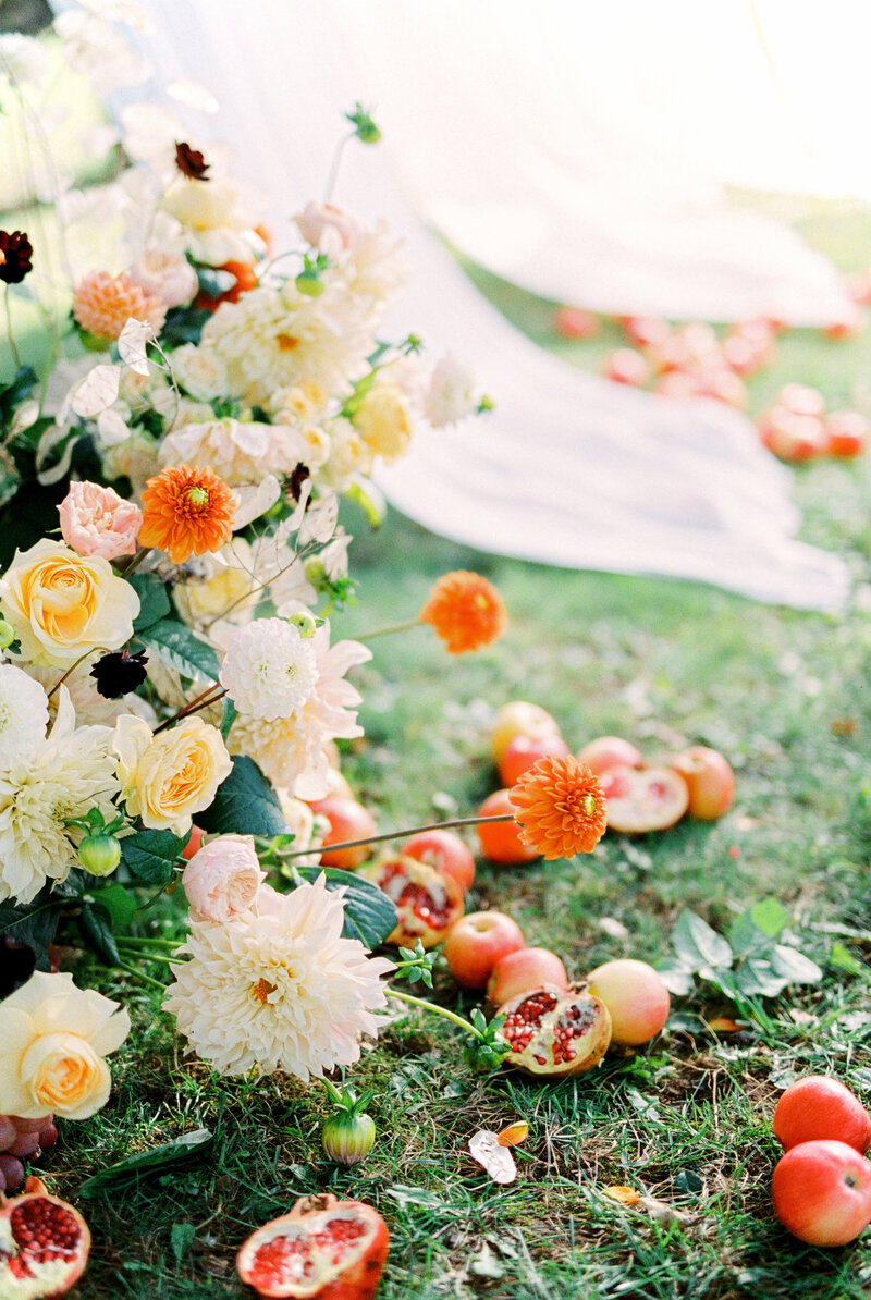 Artistic wedding flatlay with orange and cream flowers, pomegranates, and scattered fabric on grass, captured in fine art style