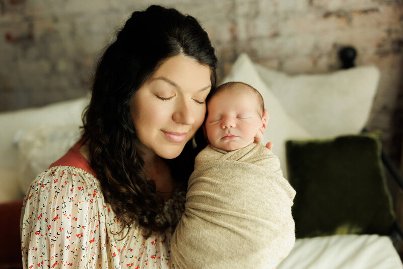 baby swaddled in a tan swaddle mom in a floral dress snuggled into her newborn baby with her eyes closed