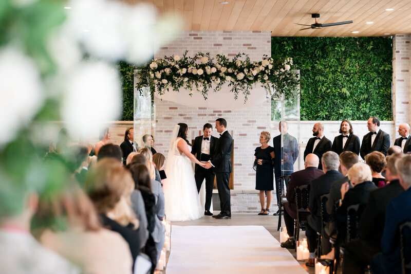 A bride and groom hold hands at the altar during their indoor wedding ceremony, surrounded by guests and floral decorations at Grbic Events, a wedding venue in St. Louis.