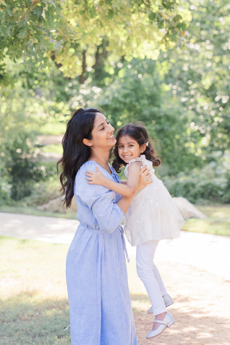 A mother dressed in a long blue dress holds her little girl on a sandy path with trees in the background at Katherine Fleischer Park. The little girl looks at the camera smiling, in this candid photo captured by an Austin Family Photographer
