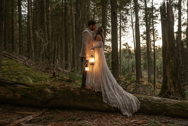 A bride and groom share an intimate moment on a fallen tree holding lanterns in a quiet forest at dusk, captured by Sydney Breann Photography during their Glacier National Park elopement.