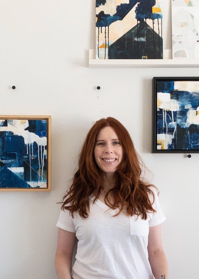 Girl with red hair, leaning against wall with hands in pockets, surrounded by art.