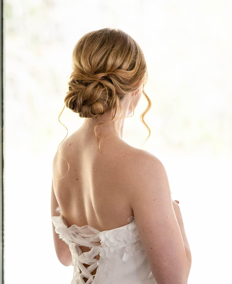 Bride in white gown with back to the camera to highlight her strawberry blonde hair in an updo chignon.  Dress is strapless but with a wide laced up back.
