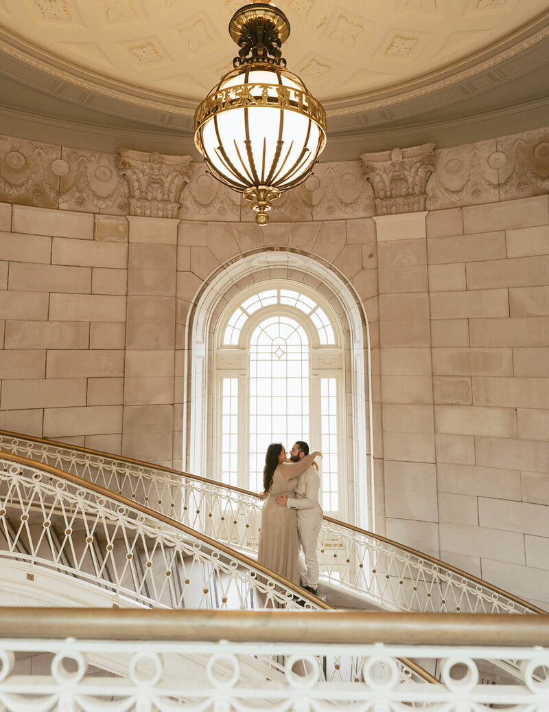 The groom and bride are hugging and kissing while standing in the middle of the staircase.