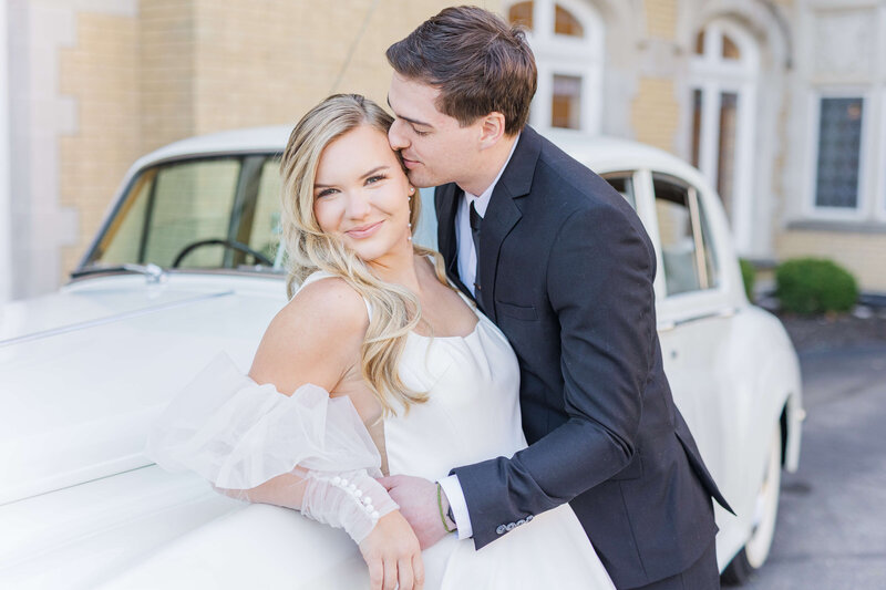 A bride and groom lean up against a car. She is smiling at the camera and he is kissing her temple.